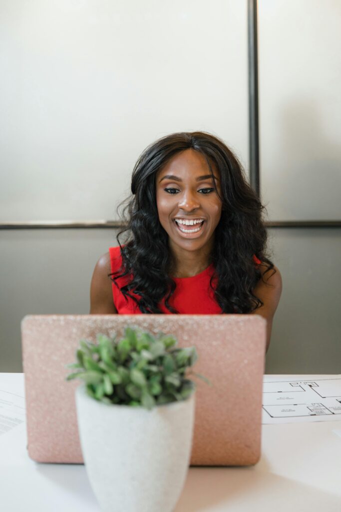 Professional woman in office, wearing red, laughing while using a laptop behind a plant.