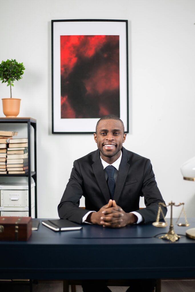 African American businessman smiling in an office environment, seated at a desk.